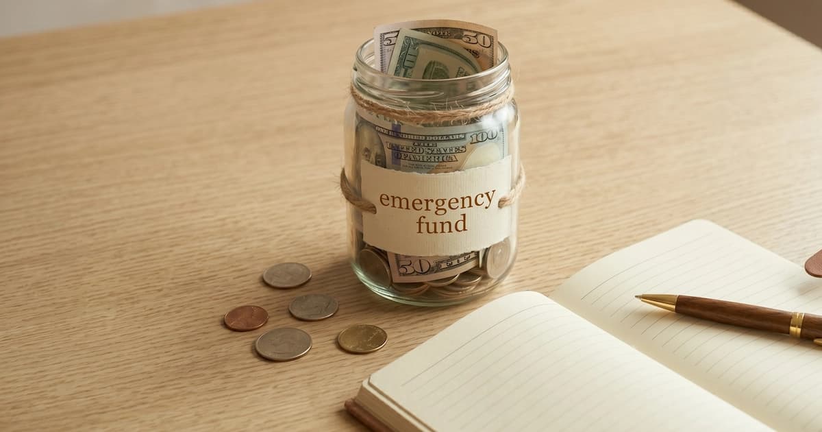 A glass jar labelled 'emergency fund' with cash inside, sitting on a wooden table